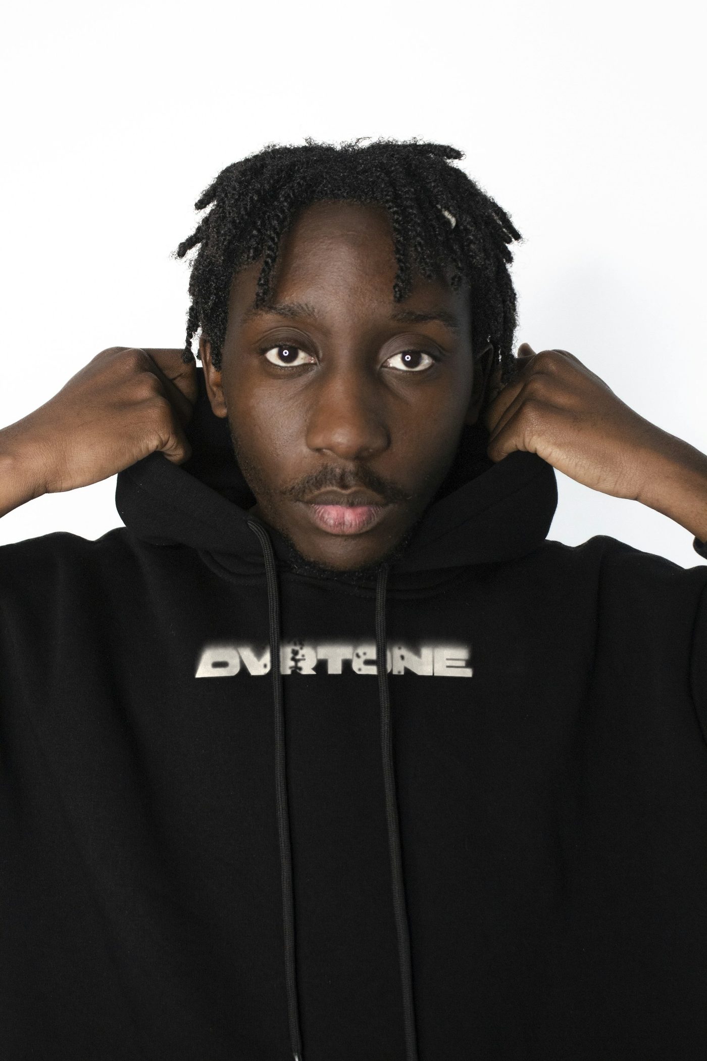 A young man pulling up the hood of a black hoodie, the OVRTONE wordmark in bone across the chest under the drawstrings