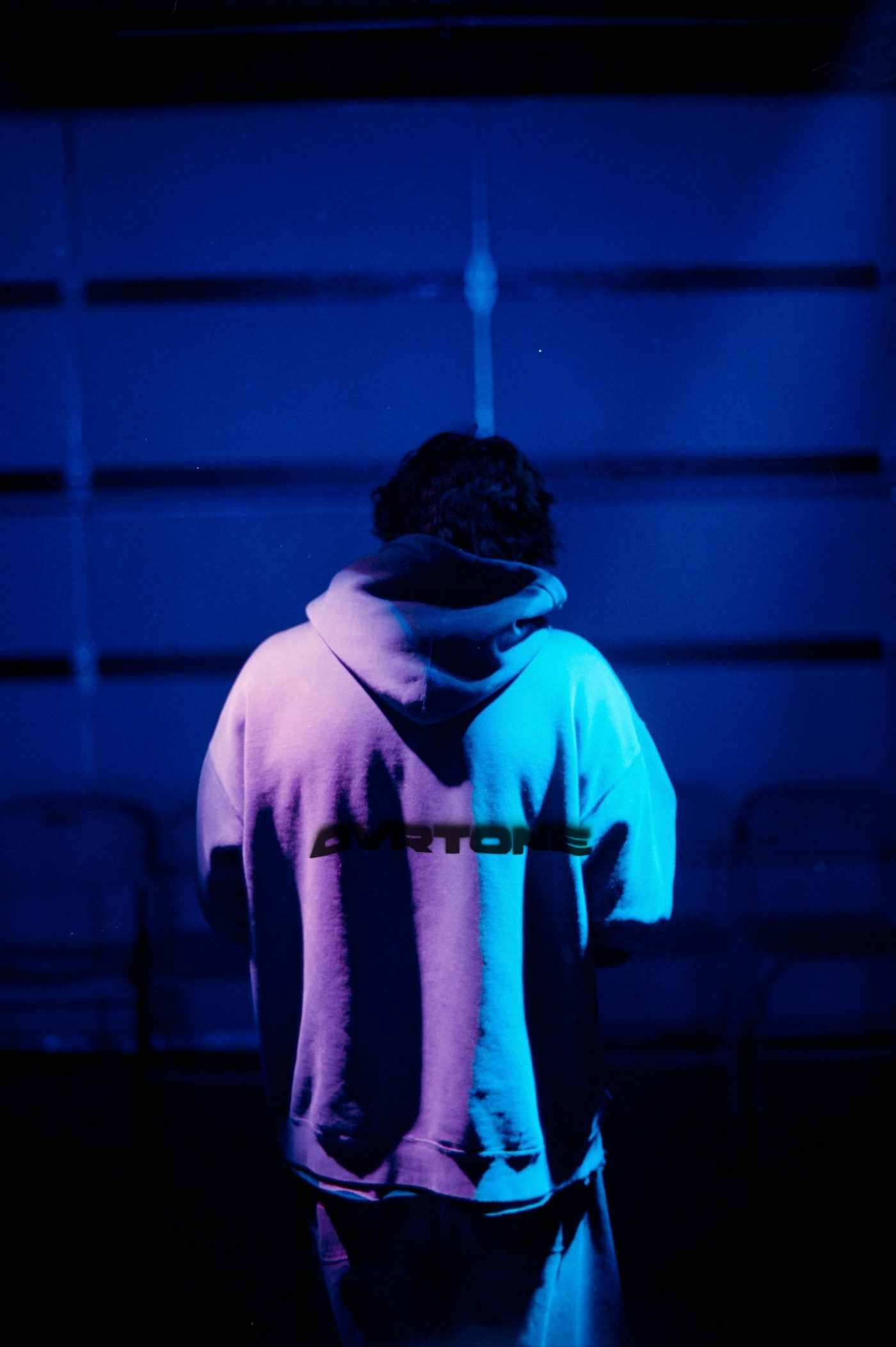 From behind, someone in an ash hoodie lit blue and purple, the OVRTONE wordmark in black across the back